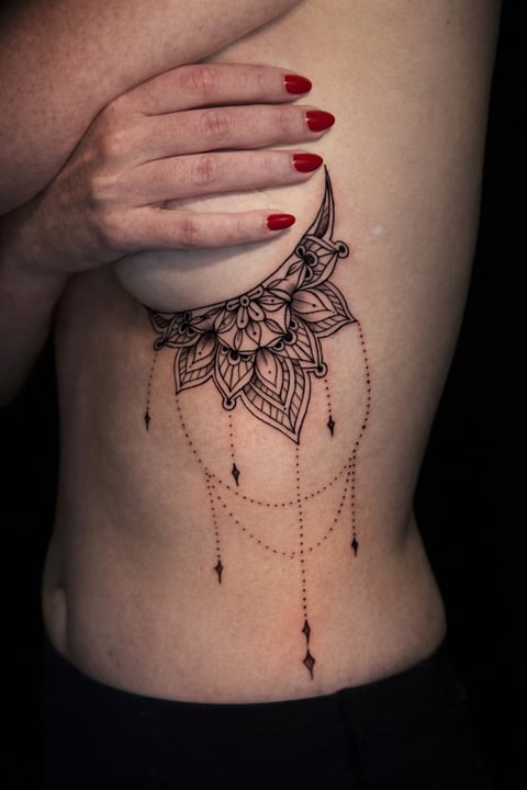 Woman with red nail polish displaying a large mandala tattoo on her ribcage with decorative hanging chains and leaf designs