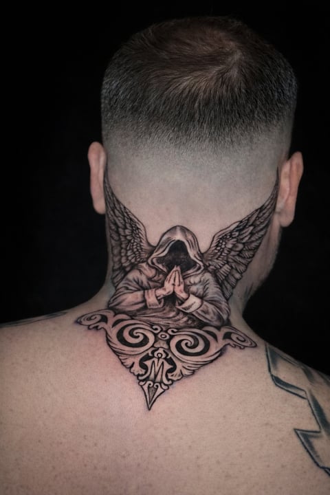 Back of mans head and neck showing a detailed black tattoo of an angel with wings and ornate scrollwork design against black background