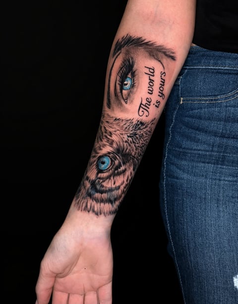 Forearm tattoo featuring a human eye with blue iris and a wolf eye with text reading The lord is yours between them on a person wearing blue jeans