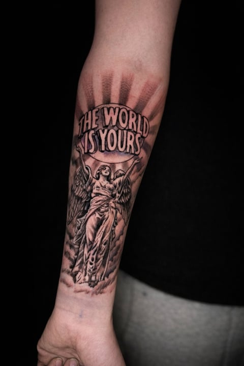 Black and gray tattoo on forearm featuring an angel statue holding a banner with text reading The World Is Yours