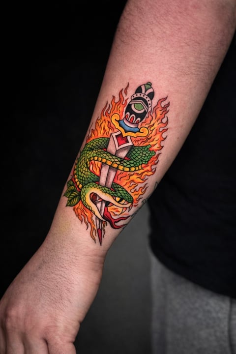 Colorful tattoo on forearm featuring a green serpent wrapped around stacked gifts with flames and a grenade on top