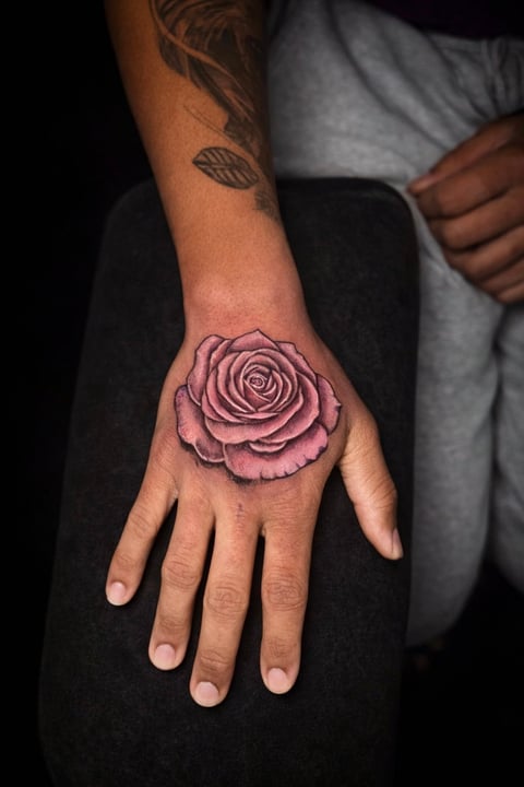 Hand tattoo of a detailed pink rose on the back of a persons hand against a dark background