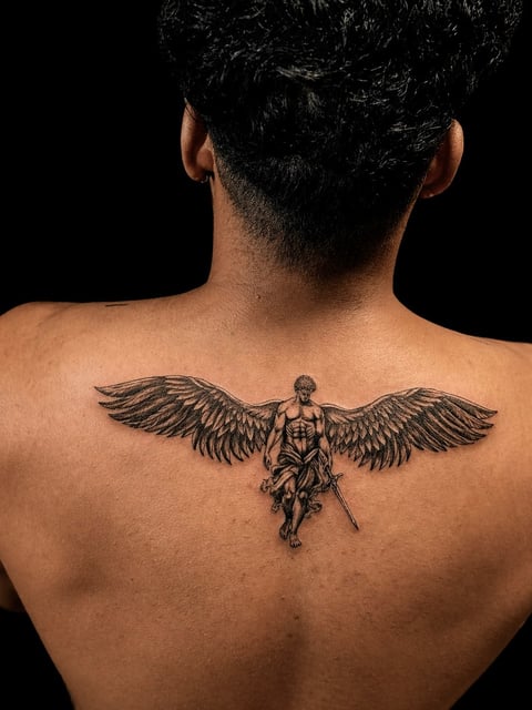 Back view of a man with a black ink tattoo of an angel with large wings spanning across his upper back against a dark background