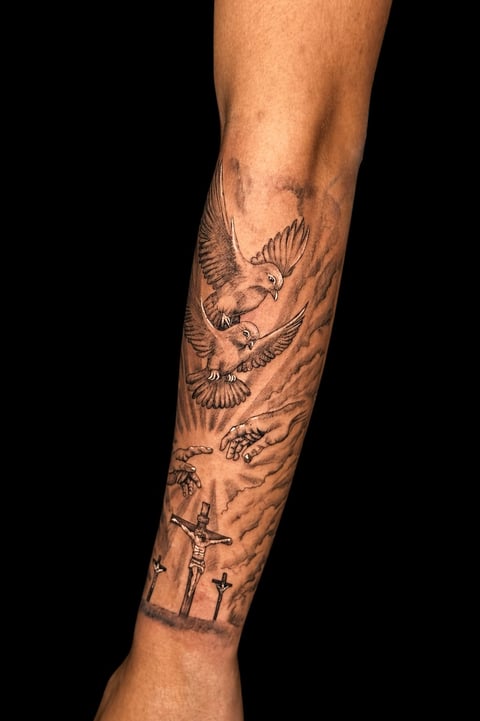 Full sleeve tattoo on forearm featuring flying doves, religious imagery including a crucifix, and decorative line work on tan skin against black background