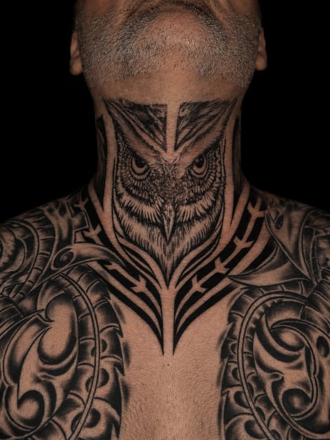Mans torso and neck with extensive black ink tattoos including an owl face design on the throat and ornamental patterns on shoulders and chest
